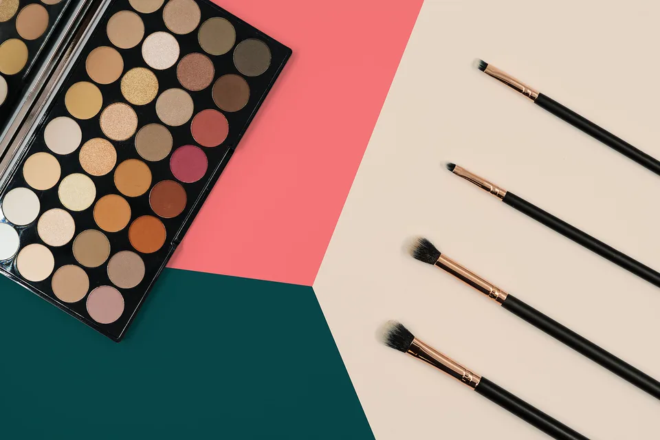 Large eyeshadow palette beside four makeup brushes on pink cream and green color blocks