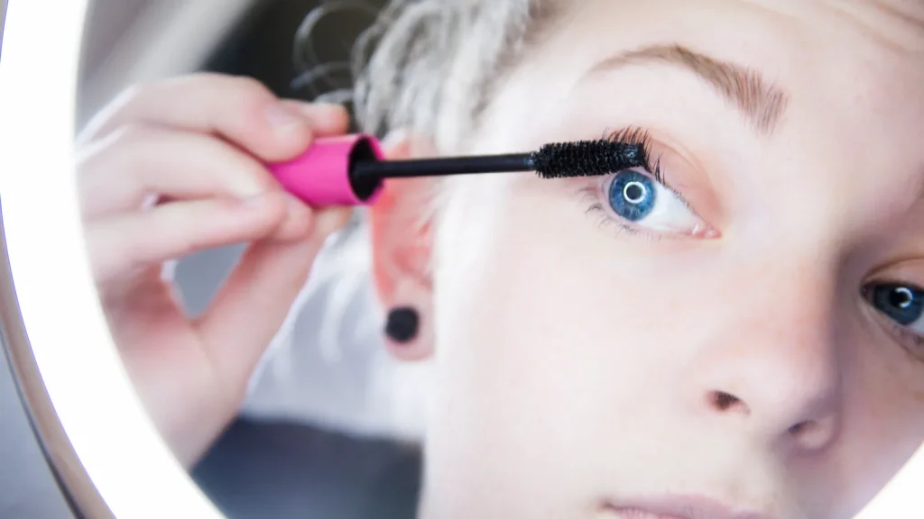 A woman applying mascara to her upper lashes in a mirror